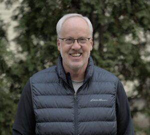 The smiling face of a white man with grey hair, a receding hairline and glasses.